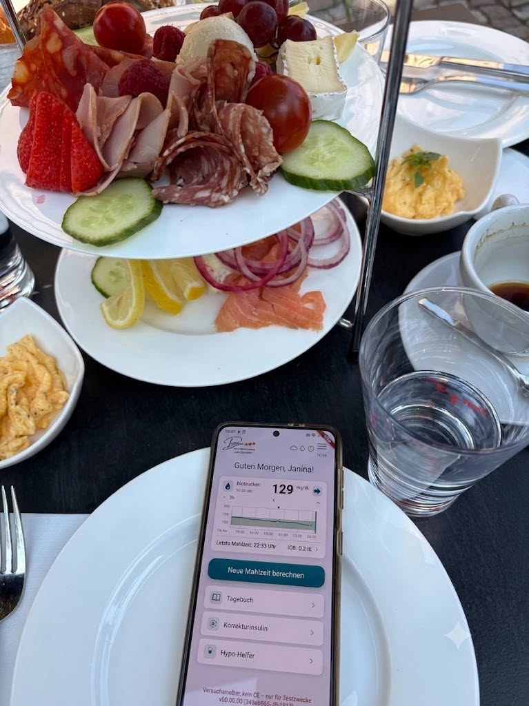 Etagere-Fruehstueck mit Aufschnitt, Kaese, Obst und Lachs; im Vordergrund der Diabakus-Startbildschirm 'Guten Morgen, Janina!' mit Blutzucker 129 mg/dl.